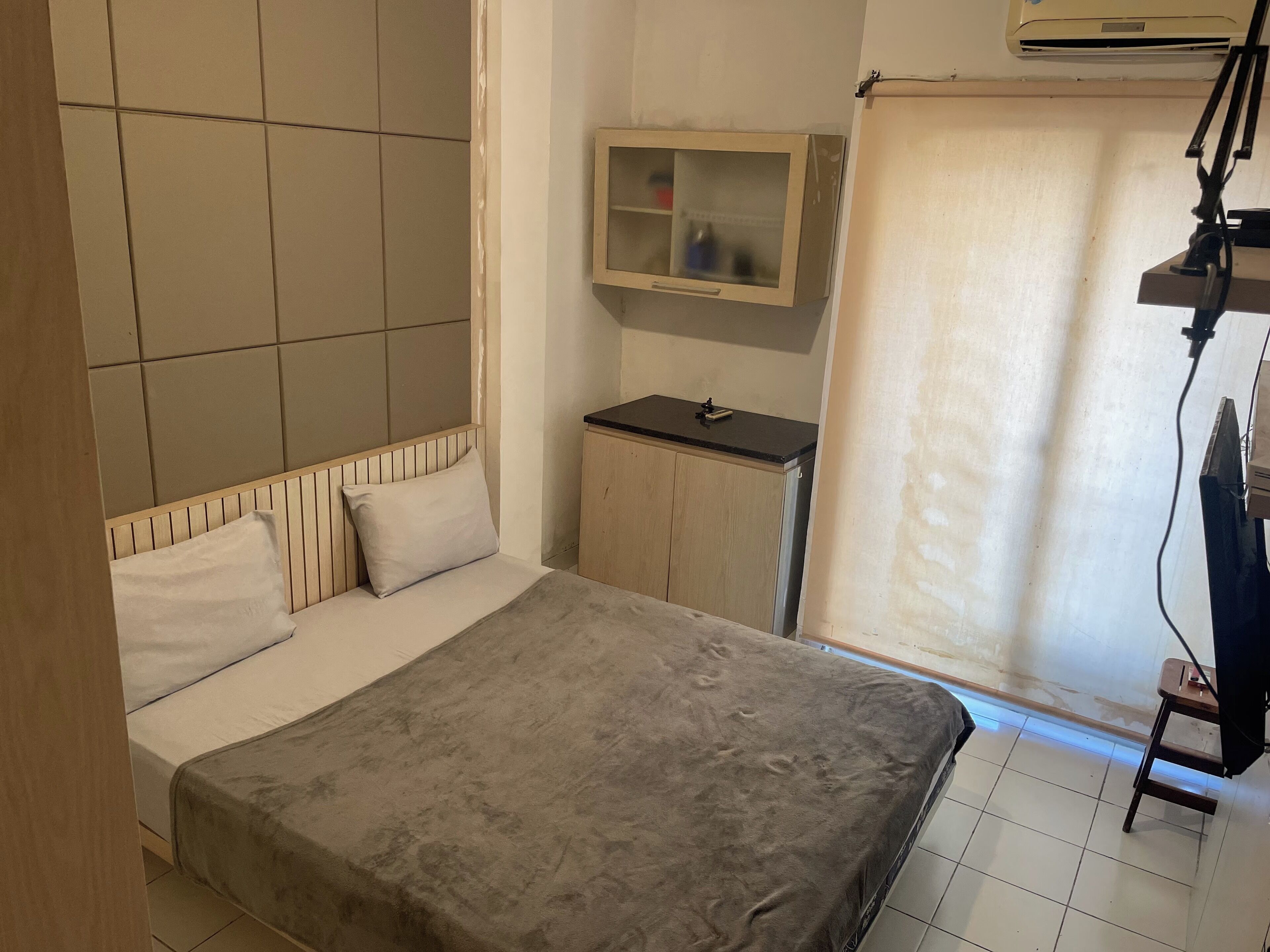 A bedroom with a bed, a small cabinet, a television, and a window with a curtain.