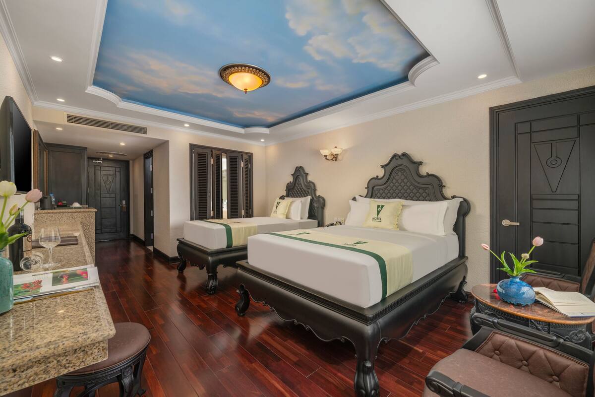 A hotel room with a bed, two bedside tables, a desk with a chair, a vanity with a mirror, and a ceiling with a painted sky design.