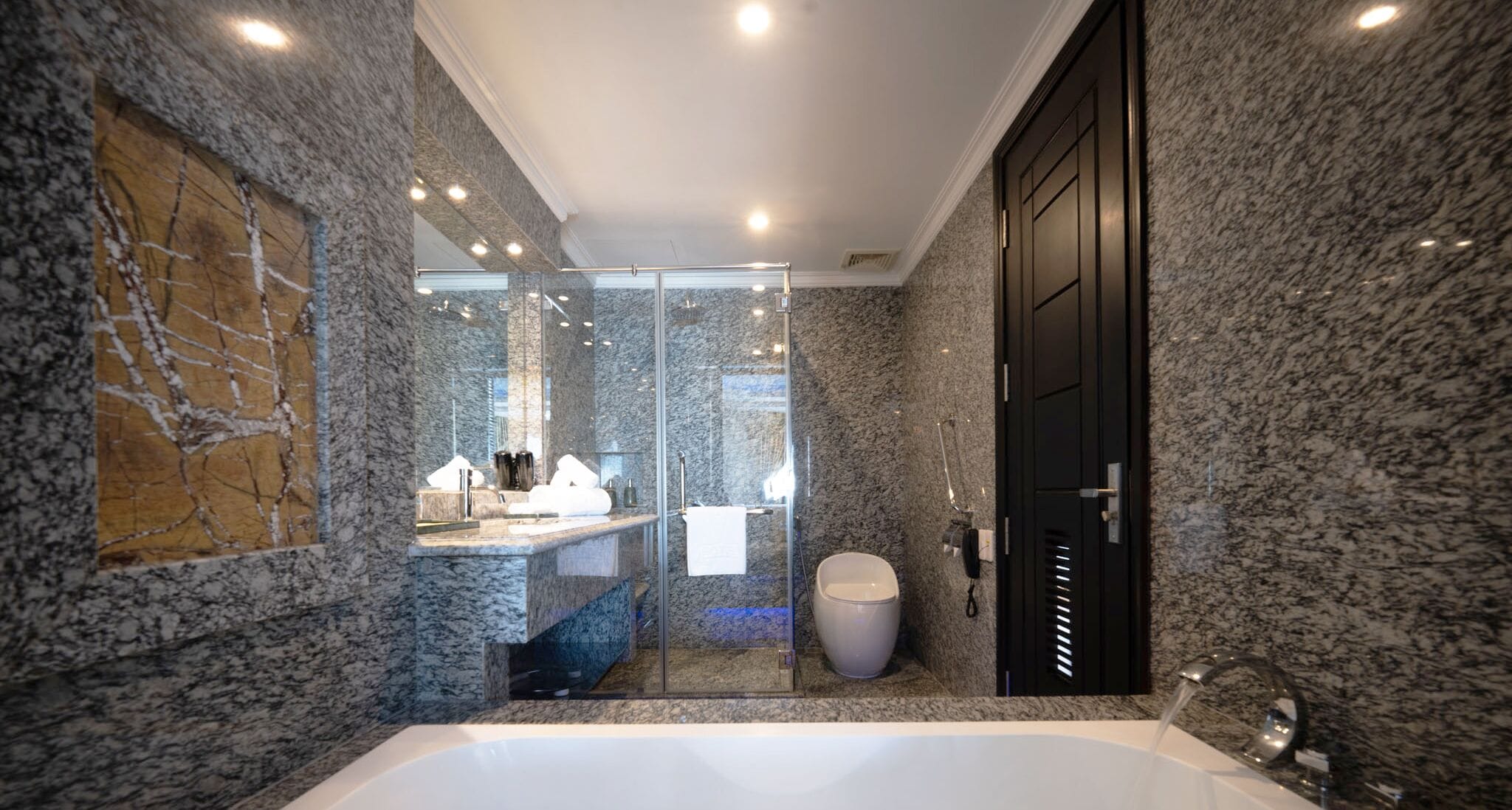 A modern bathroom with a marble wall, a large bathtub, a glass-enclosed shower, and a vanity with a mirror.