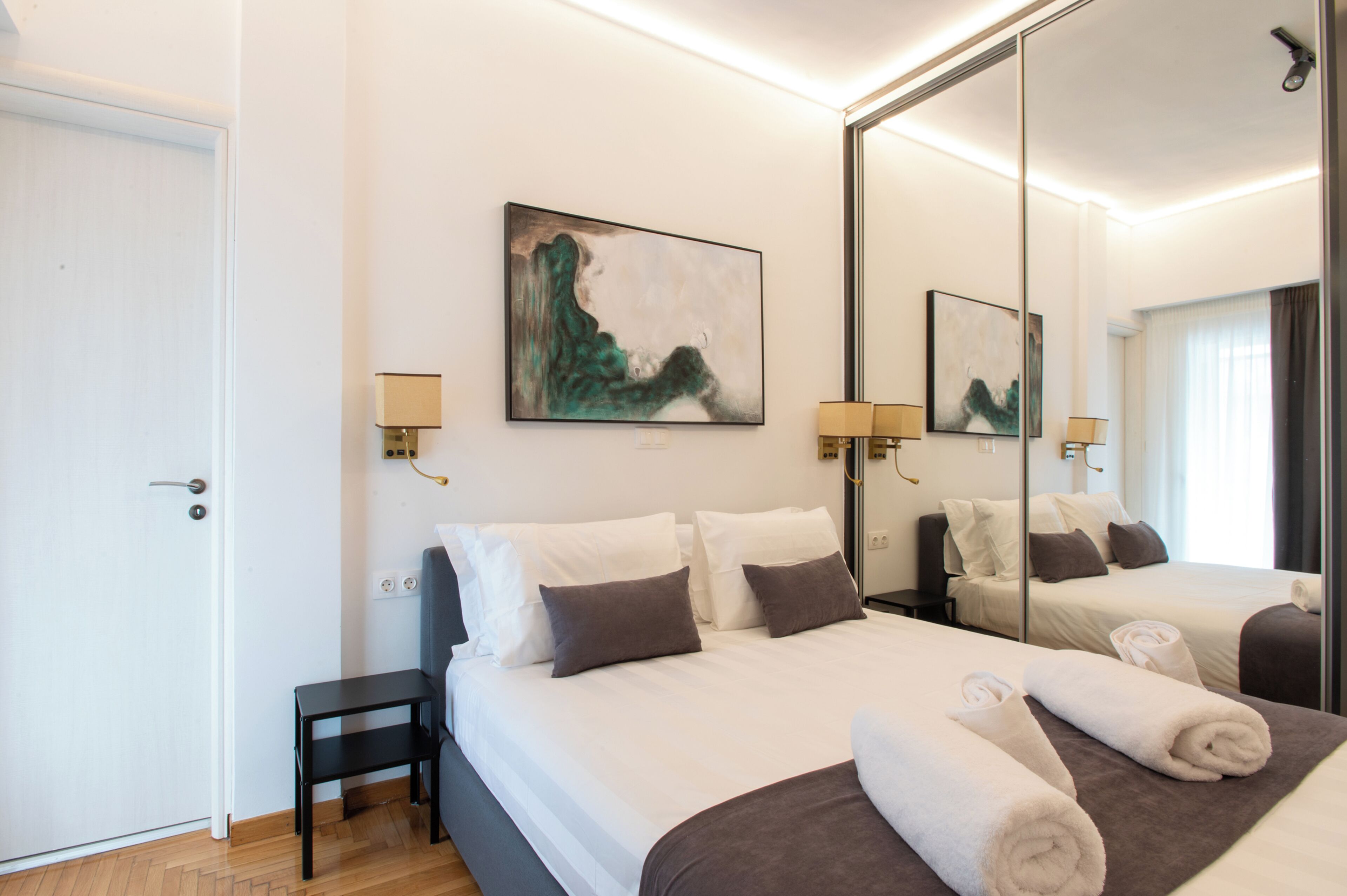 A hotel room with a large bed, a small table, a chair, a wall-mounted lamp, and a painting.
