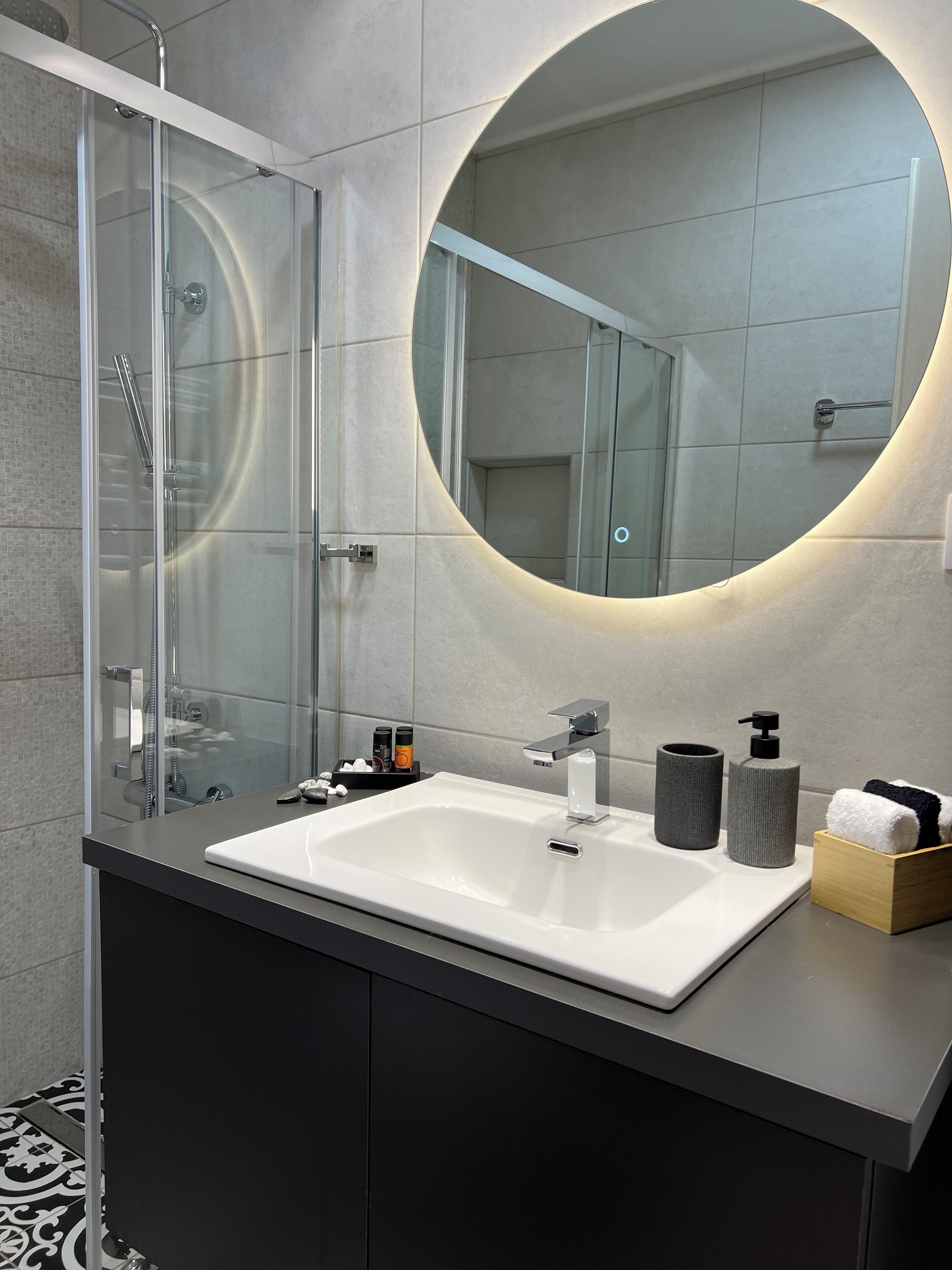 Un baño moderno con cabina de ducha de vidrio, un espejo redondo con luz y un lavamanos blanco con grifo.