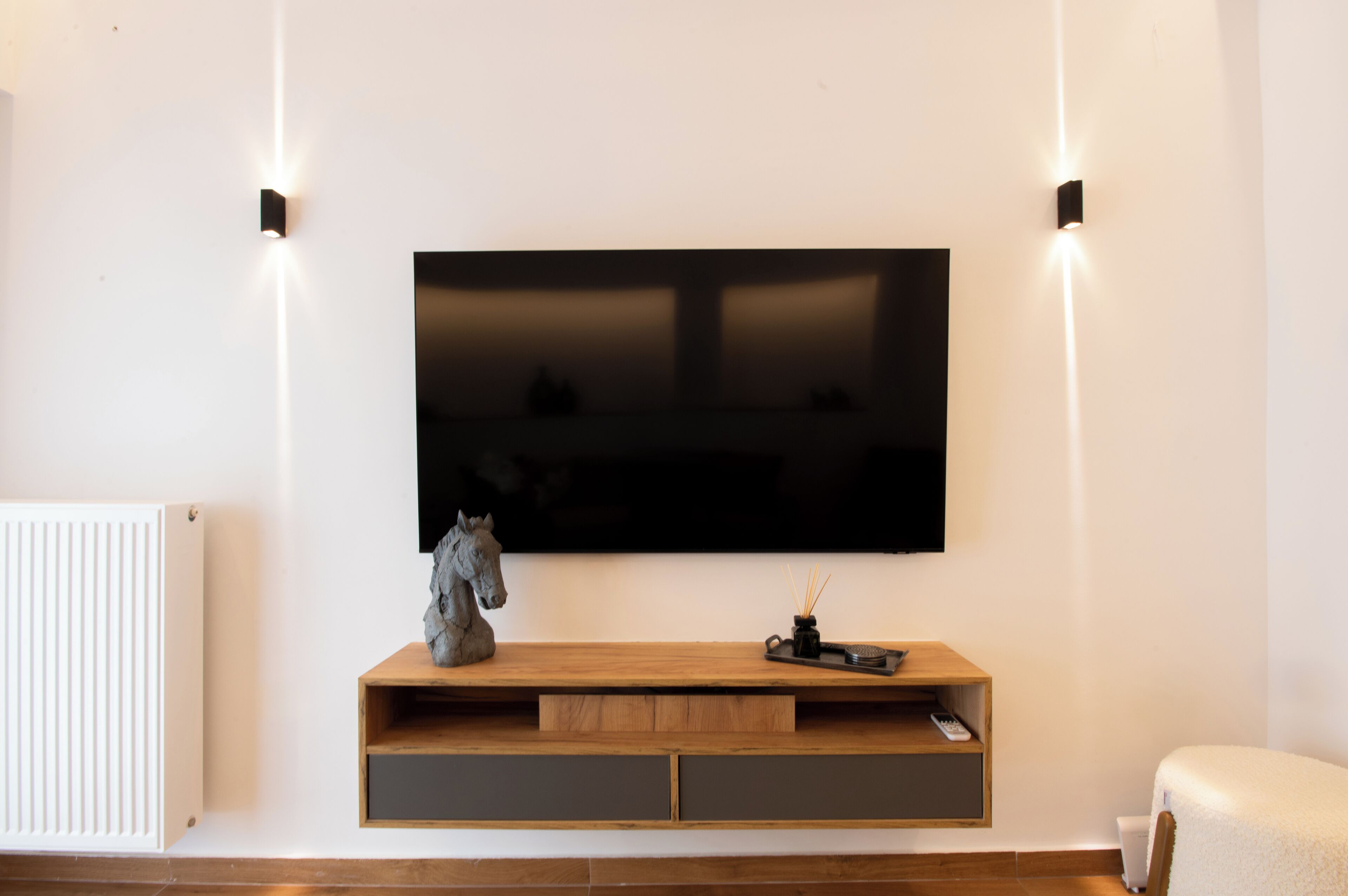 Een moderne woonkamer met een flatscreen televisie aan de muur, een houten console met een beeld van een paard, en een radiator links.