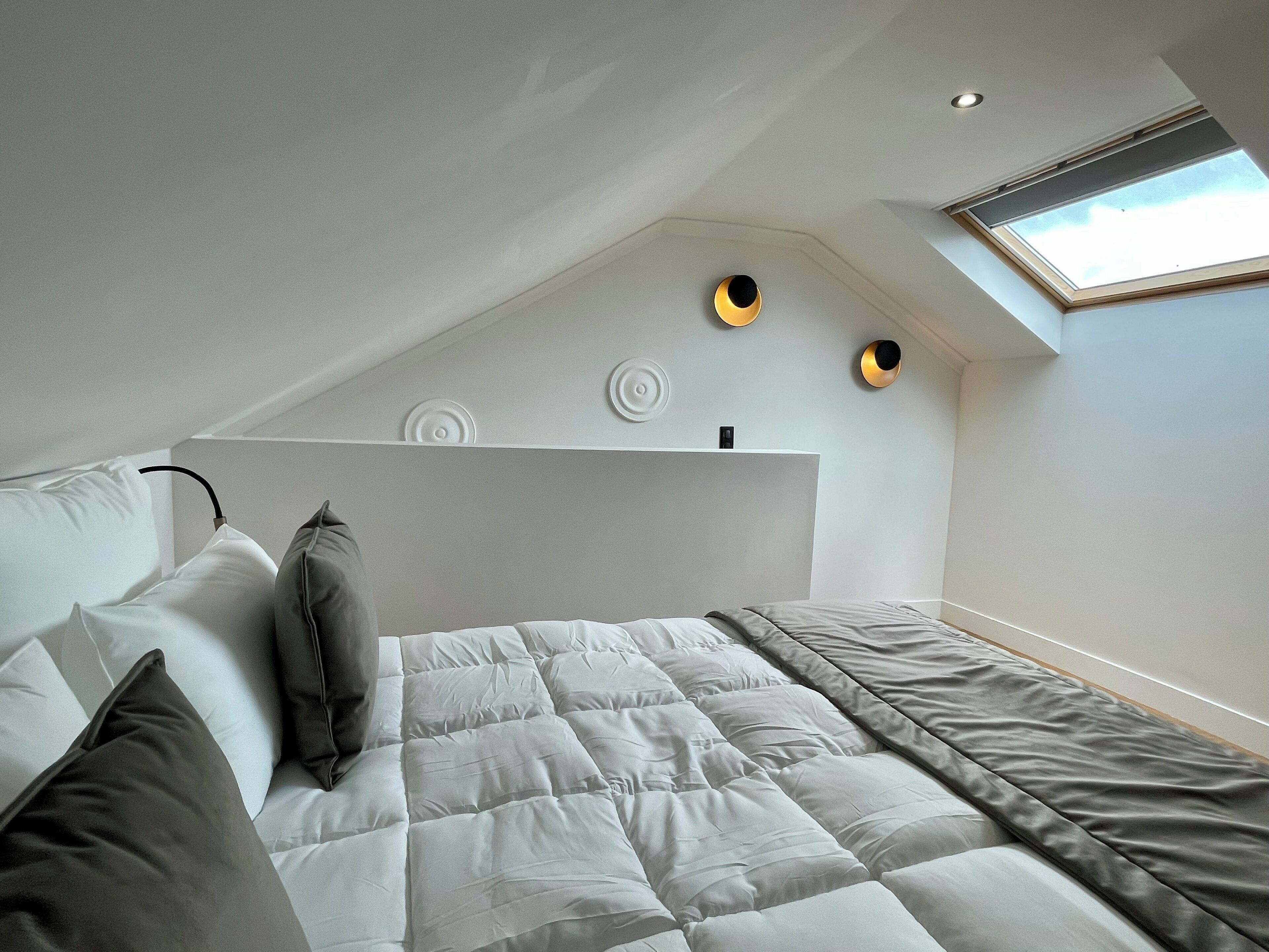 Un dormitorio con techo inclinado, tragaluz, cama con ropa de cama blanca y gris, y apliques decorativos en la pared.