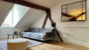 A modern loft bedroom with a sloped ceiling, a grey sofa, a round ottoman, a small table, and a floor lamp. - Alfred Suites (Dijon)