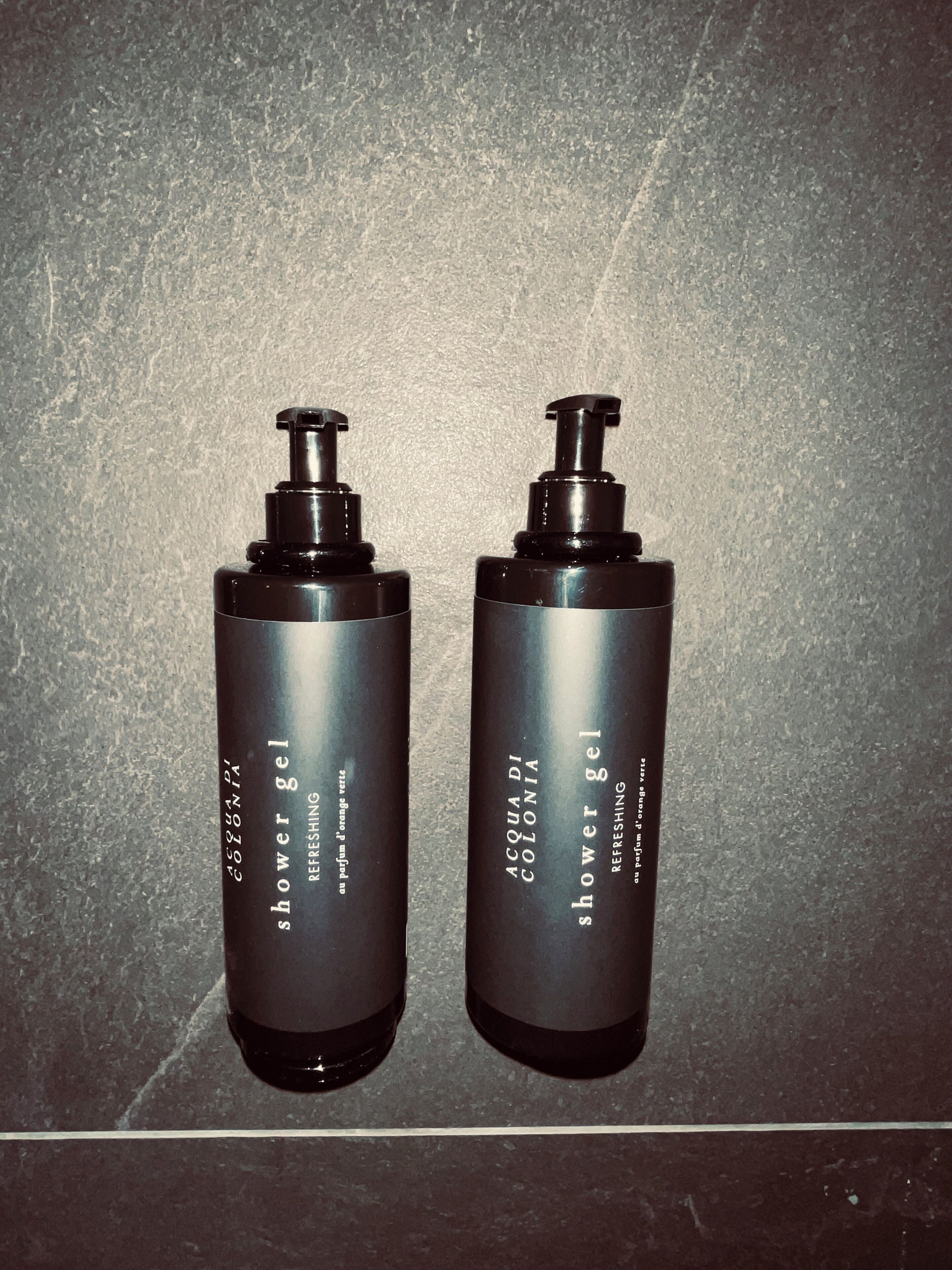 Dos botellas negras etiquetadas como 'gel de ducha', con la etiqueta 'refrescante' y 'aromático de naranja verde' sobre una superficie texturizada.