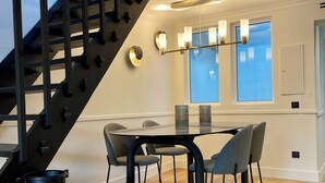 A modern dining area with a glass table, grey chairs, a black staircase, and a ceiling light fixture. - Alfred Suites (Dijon)