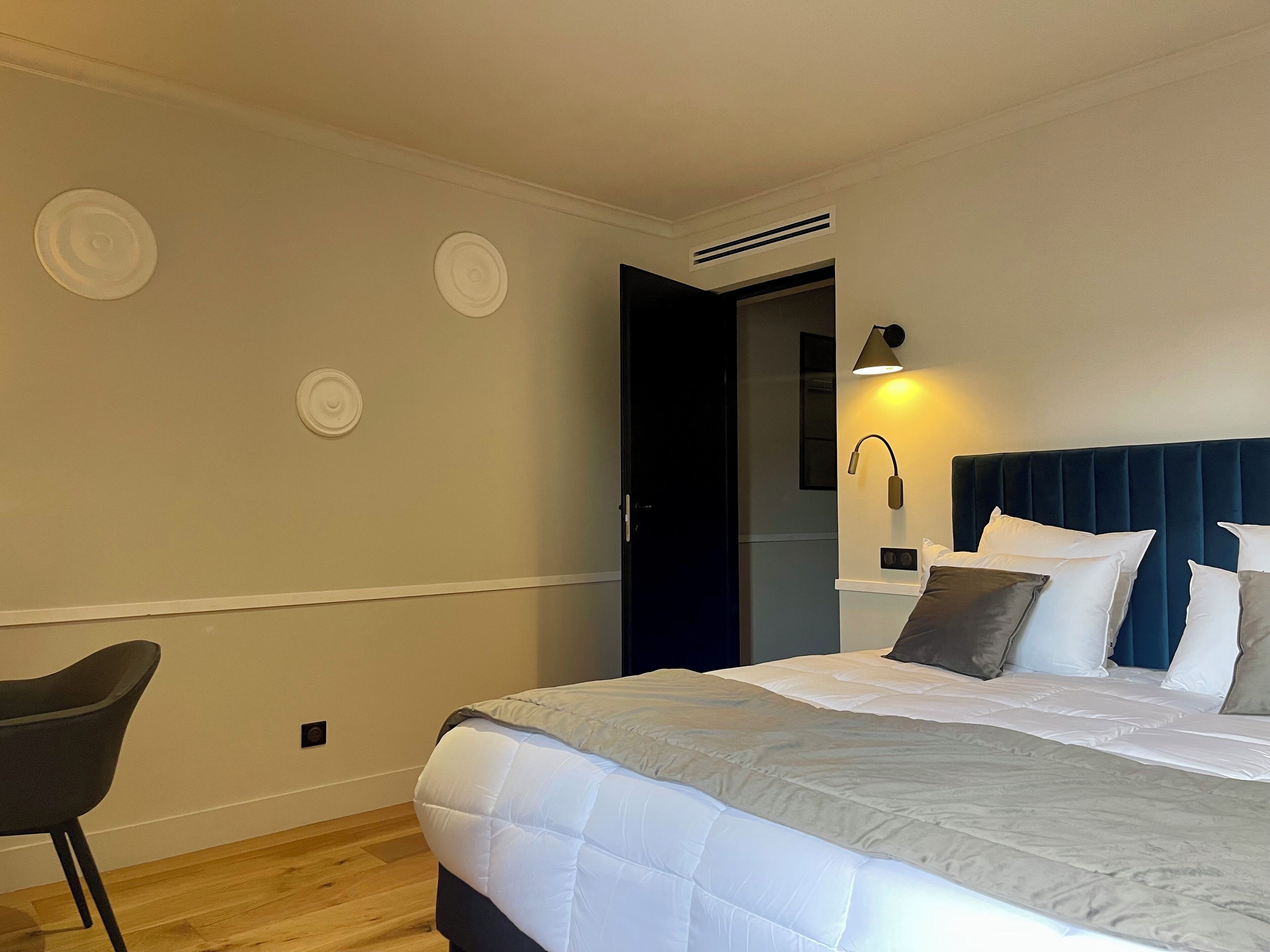 A hotel room with a bed, a chair, a bedside table, a lamp, and a door leading to another room.