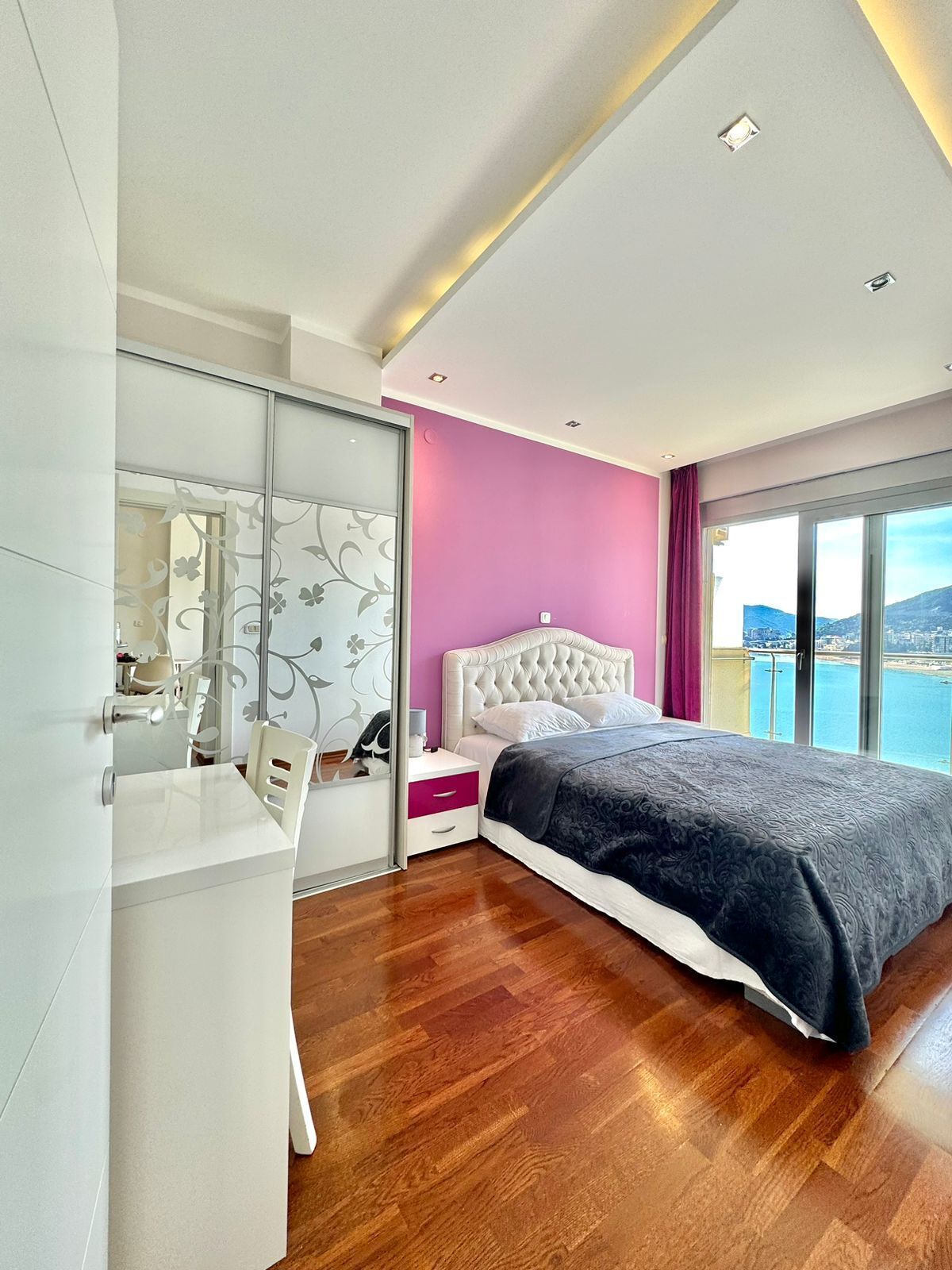 A modern bedroom with a large bed, a vanity with a mirror, and a view of the sea.