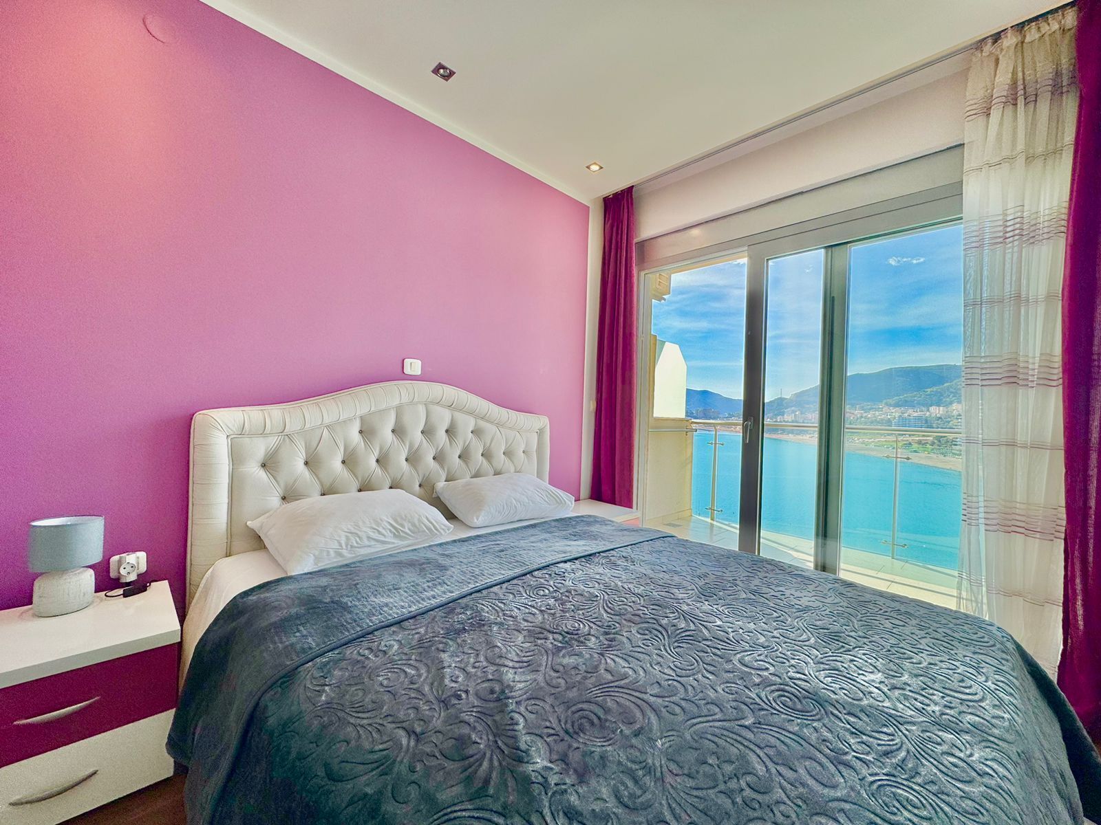 A bedroom with a large bed, a nightstand, a window with a view of a body of water and mountains, and a pink wall.