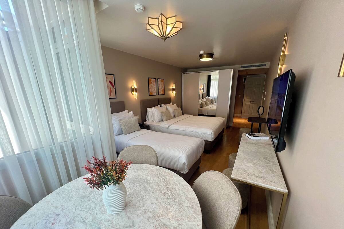 Habitación de hotel con dos camas, una mesa de comedor con sillas, un jarrón con flores y un televisor.