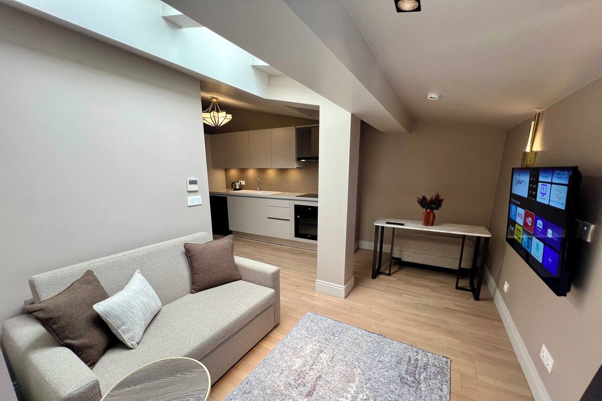 Una sala moderna con un sofá, una mesa de centro y una alfombra. Se aprecia una cocina al fondo con armarios y un comedor con una mesa y sillas.