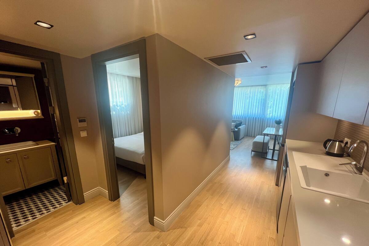 Habitación de hotel con piso de madera, una cama, baño con lavamanos y tetera, y zona de comedor con mesa y sillas.