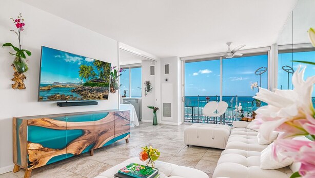 Ein modernes Wohnzimmer mit einem großen Fernseher, einem Holzkasten, einem weißen Sofa und Blick auf das Meer durch große Fenster.