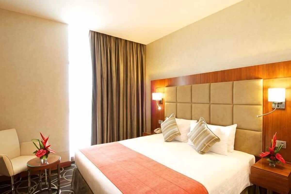 Una habitación de hotel con cama, una silla, una mesita con un florero y una mesita de noche con lámpara.