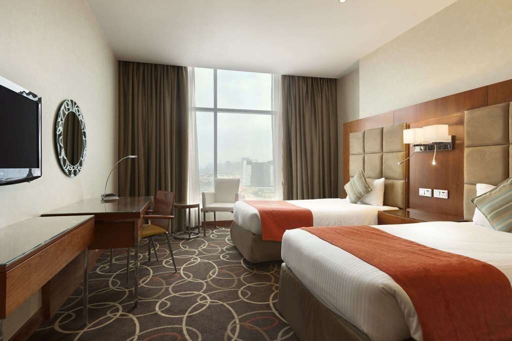 A hotel room with two beds, a desk, a chair, a TV, and a large window with curtains.