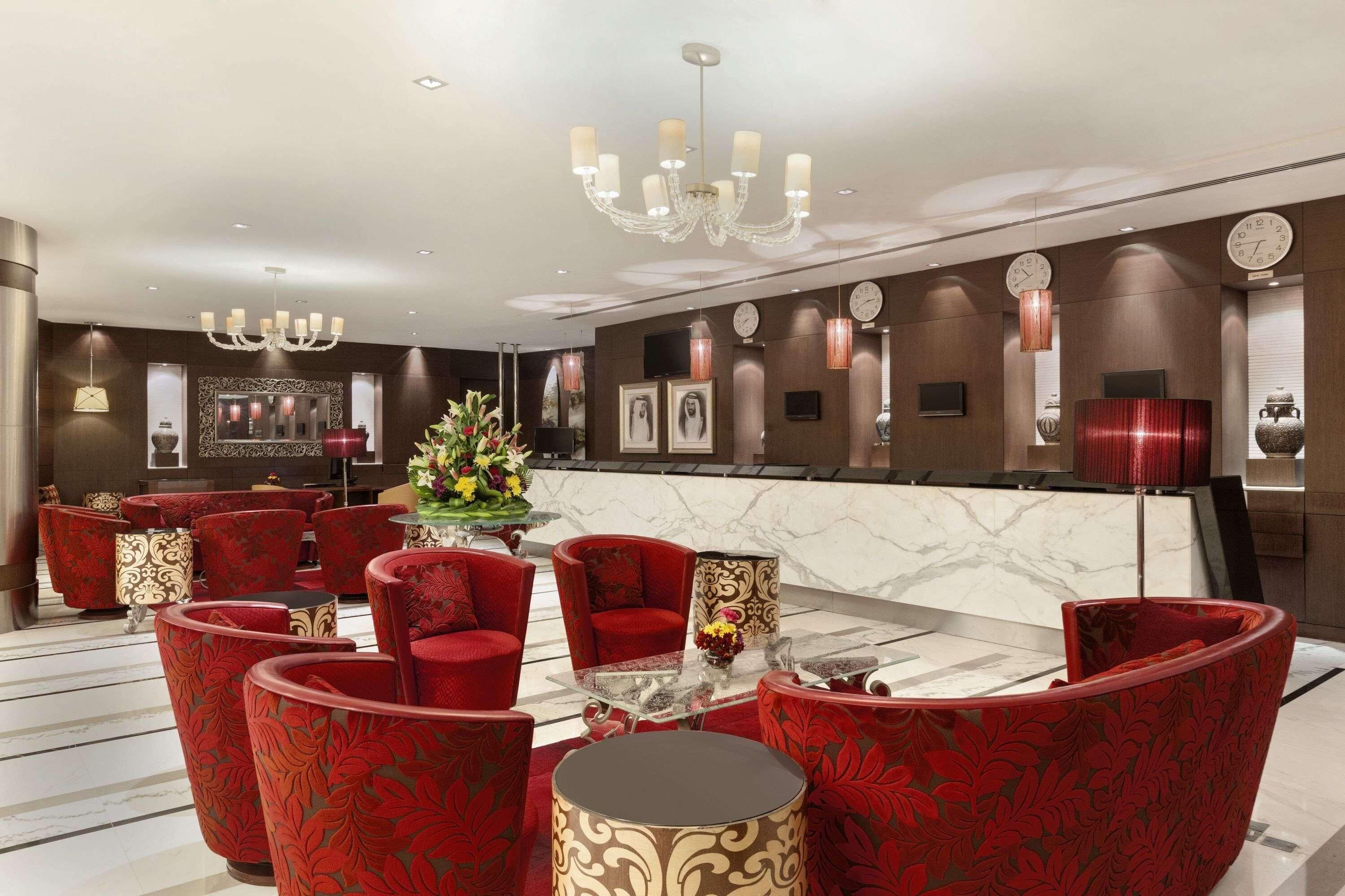 A hotel lobby with red upholstered chairs, a glass coffee table, a chandelier, and a reception desk.