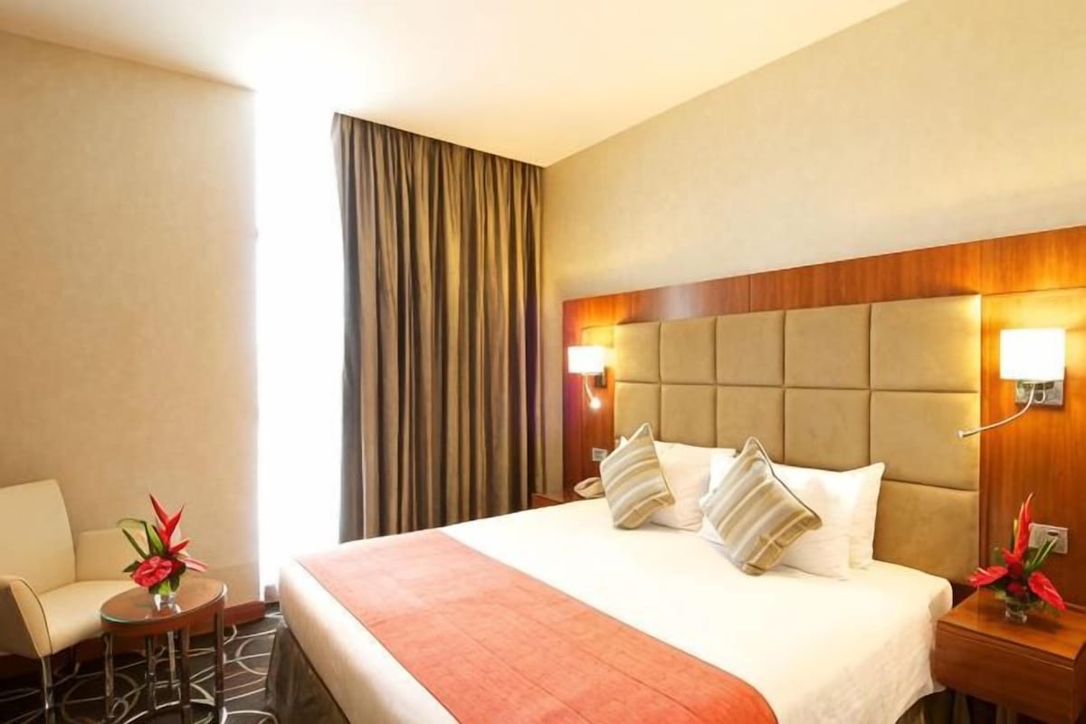Une chambre d’hôtel avec un lit, une chaise, une petite table sur laquelle se trouve un vase, et une table de chevet avec une lampe.