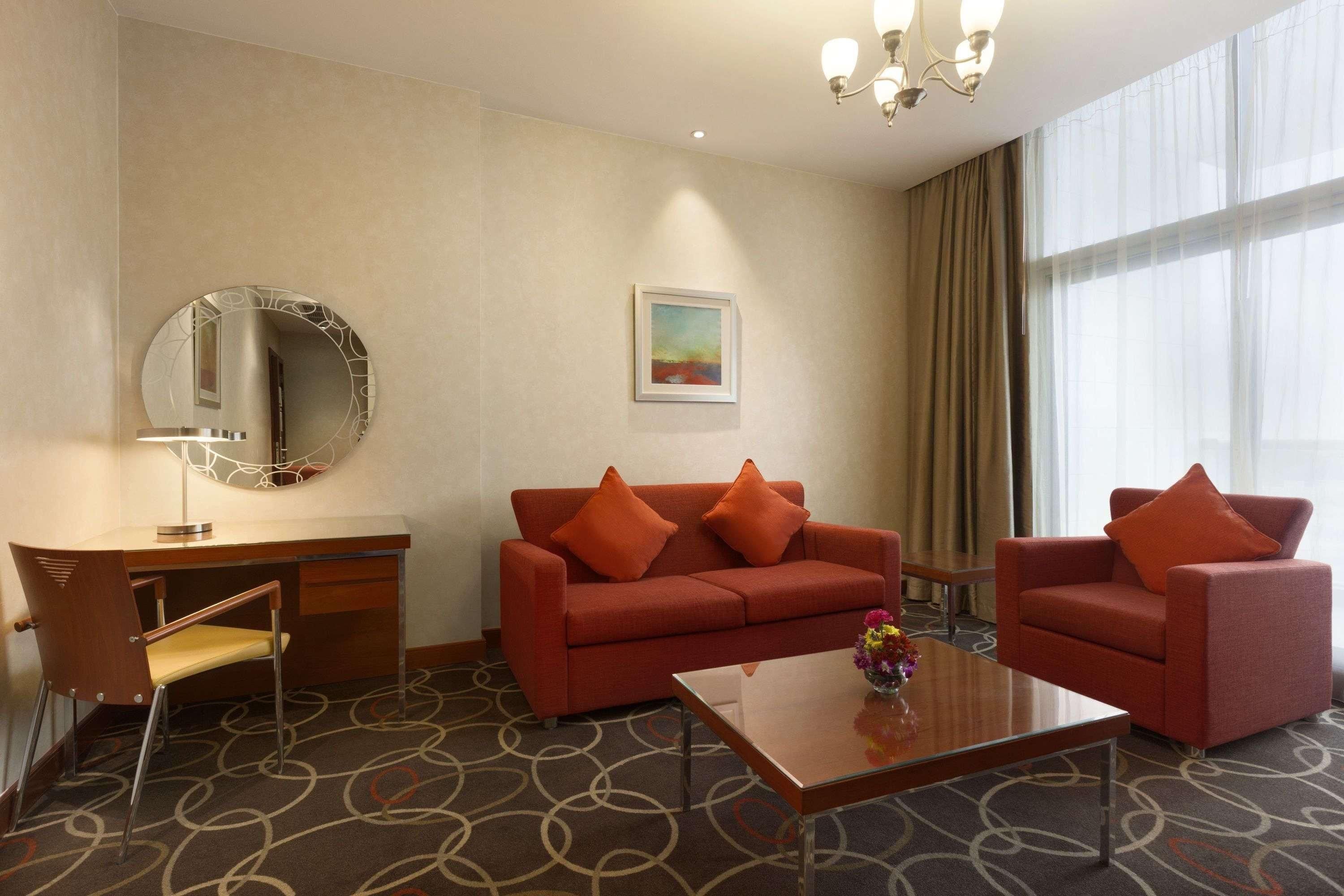A hotel room with a sofa, armchairs, a desk, a chair, a mirror, a painting, and a large window.