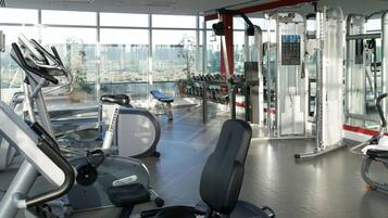 A gym with various exercise equipment, including treadmills and weight machines, with a city view through large windows.