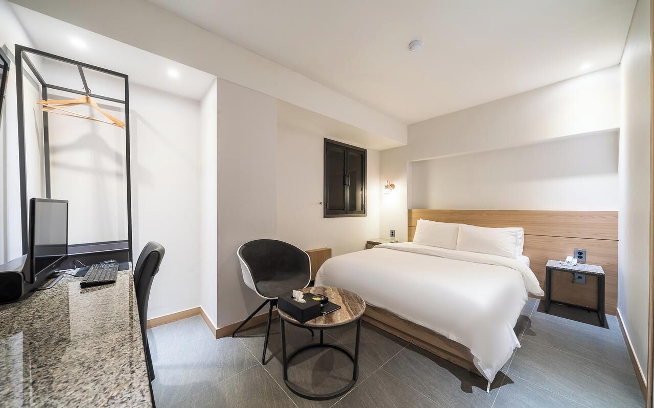 A hotel room with a bed, a desk with a chair, a small table, a TV, and a wooden headboard.