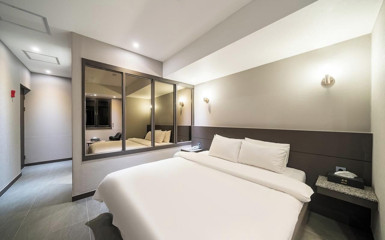 A modern hotel room with a large bed, a bedside table, a nightstand, a mirror, and a wall-mounted light fixture.