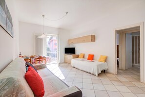 A modern hotel room with a sofa, a bed with orange pillows, a TV, and a balcony with a table and chairs. - Casa al Cor Apartments (Nago-Torbole)