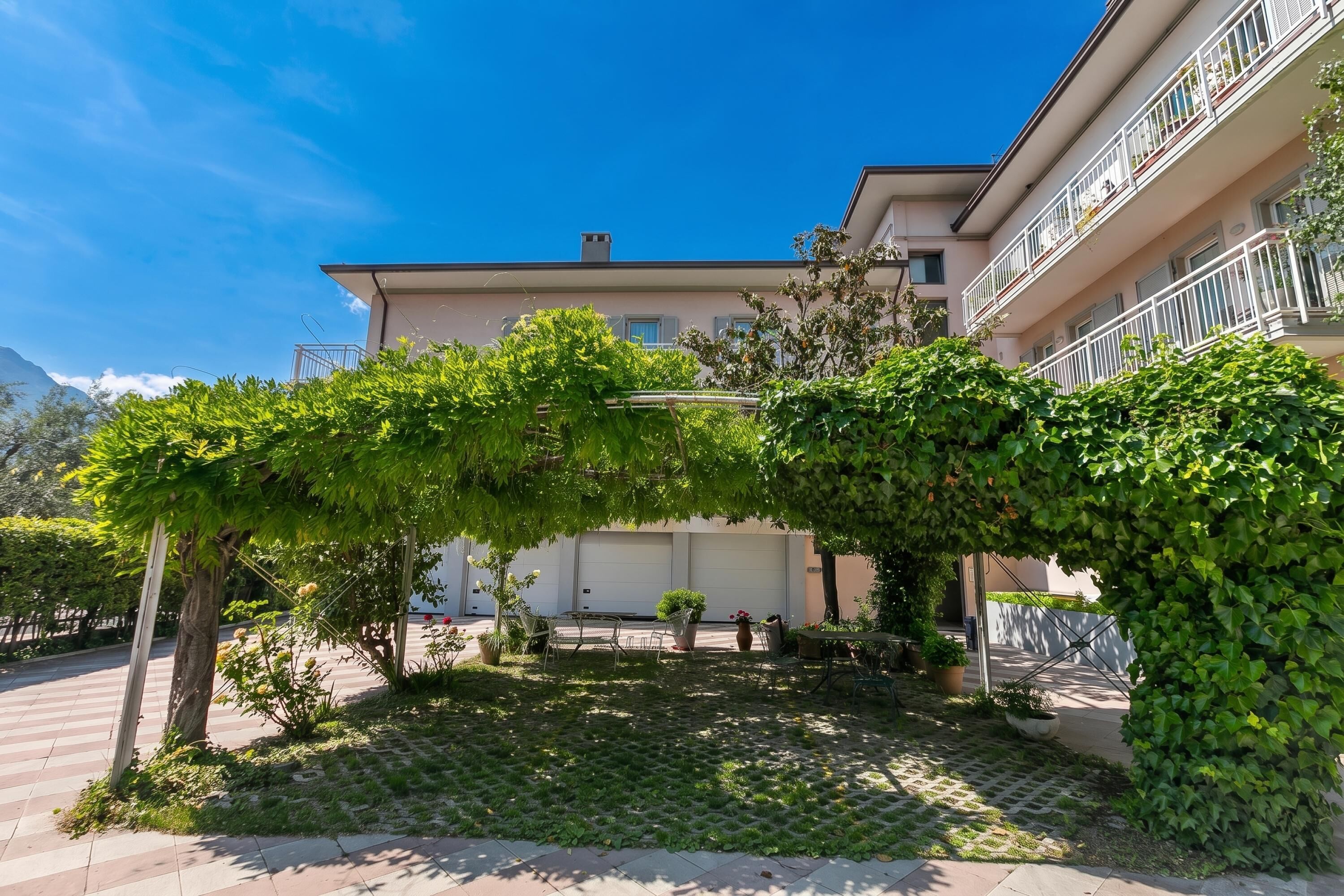 Un patio con una pérgola cubierta de follaje, un sendero empedrado y un edificio con balcones.