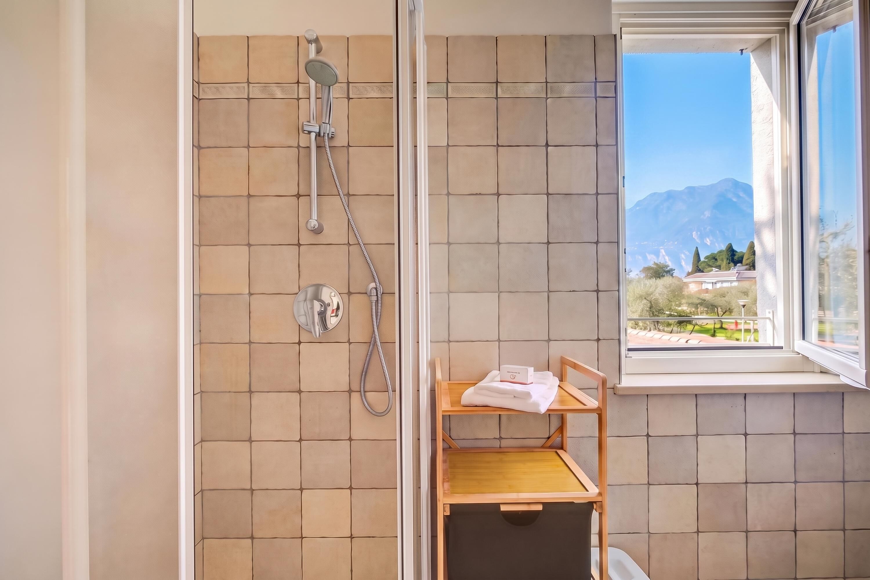 A bathroom with a shower, a window with a view of mountains, and a shelf with towels.