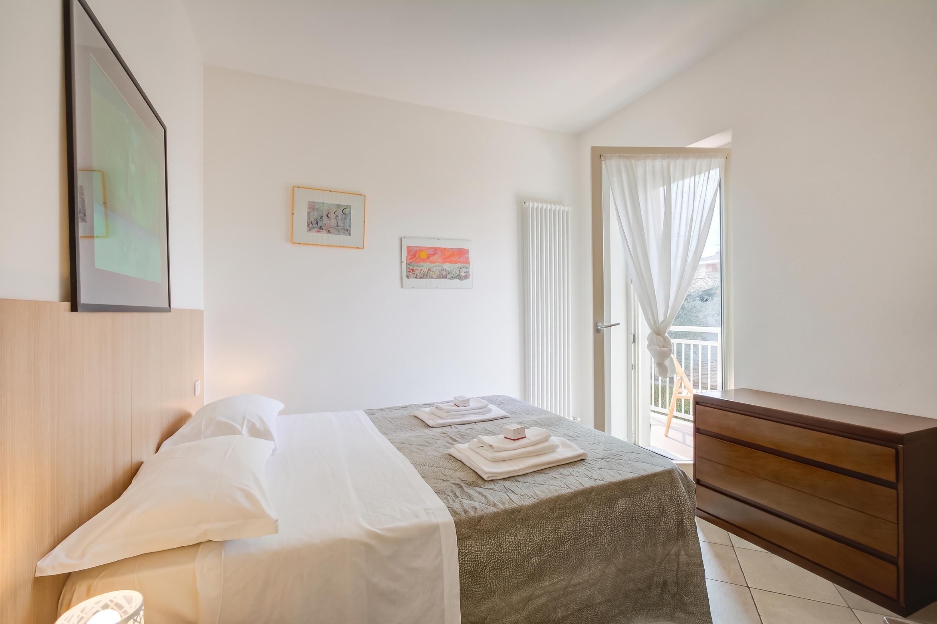 A hotel room with a bed, a wooden dresser, a window with curtains, and artwork on the wall.