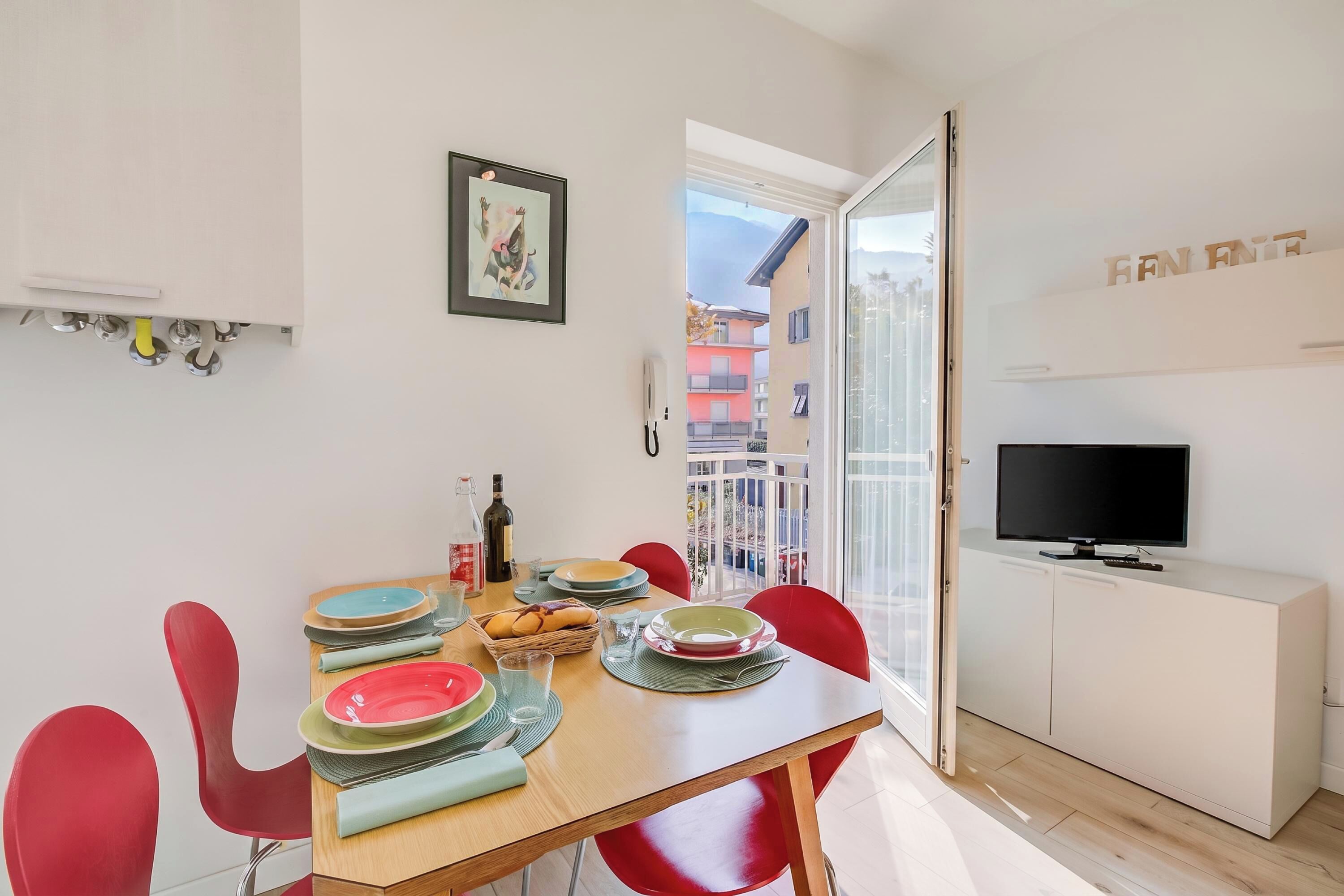 A dining area with a table set for four, red chairs, a television on a white cabinet, and a balcony with a view of buildings.