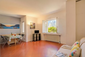 A room with a dining table, chairs, a TV on a stand, a radiator, and a window with a view of greenery. - Casa al Cor Apartments (Nago-Torbole)