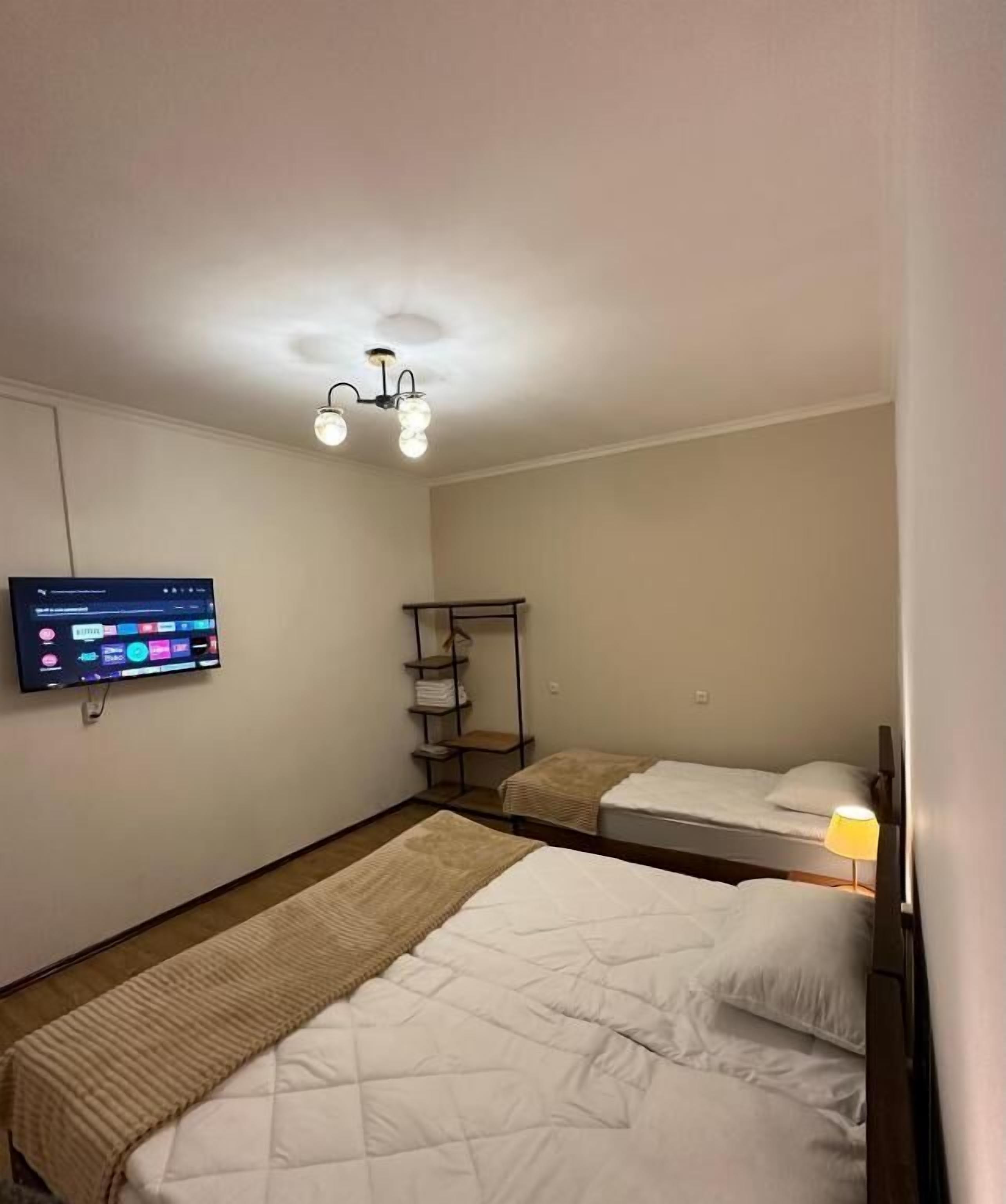 A hotel room with two beds, a TV mounted on the wall, a shelf with folded towels, and a ceiling light fixture.