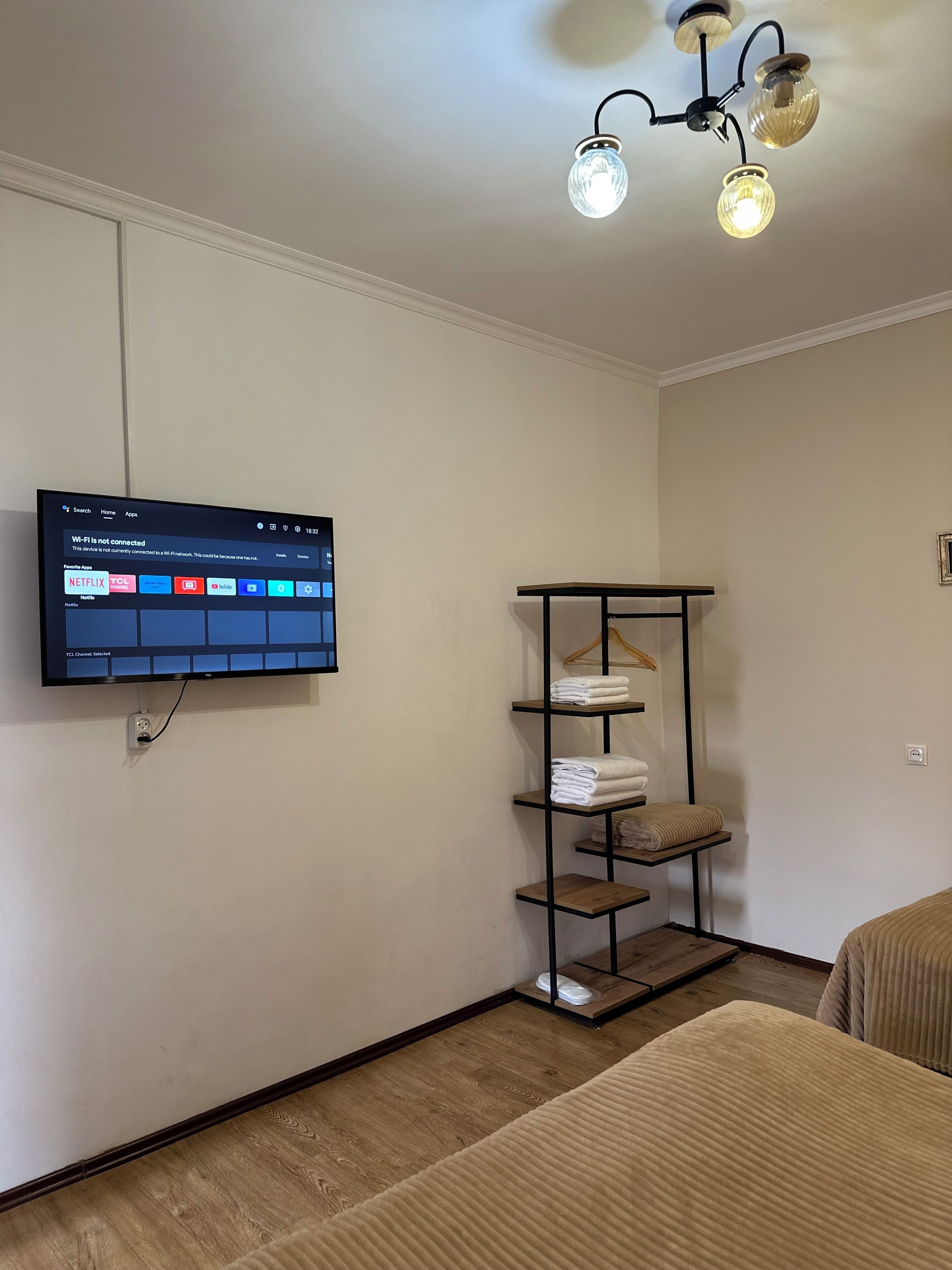 Una habitación de hotel con una televisión de pantalla plana montada en la pared, piso de madera, una cama con cubrecama color beige, un estante metálico con toallas dobladas y una lámpara en el techo.