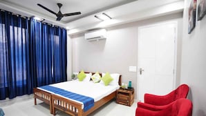 A bedroom with a wooden bed, blue curtains, a red chair, a ceiling fan, and an air conditioning unit. - Mansavi Villa HomeStay (Greater Noida)