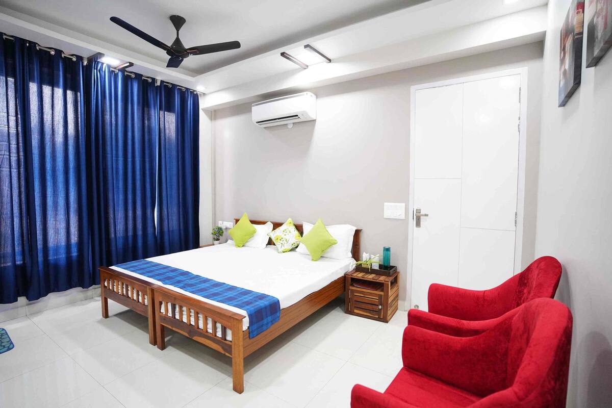Une chambre à coucher avec un lit en bois, des rideaux bleus, une chaise rouge, un ventilateur de plafond et un appareil de climatisation.