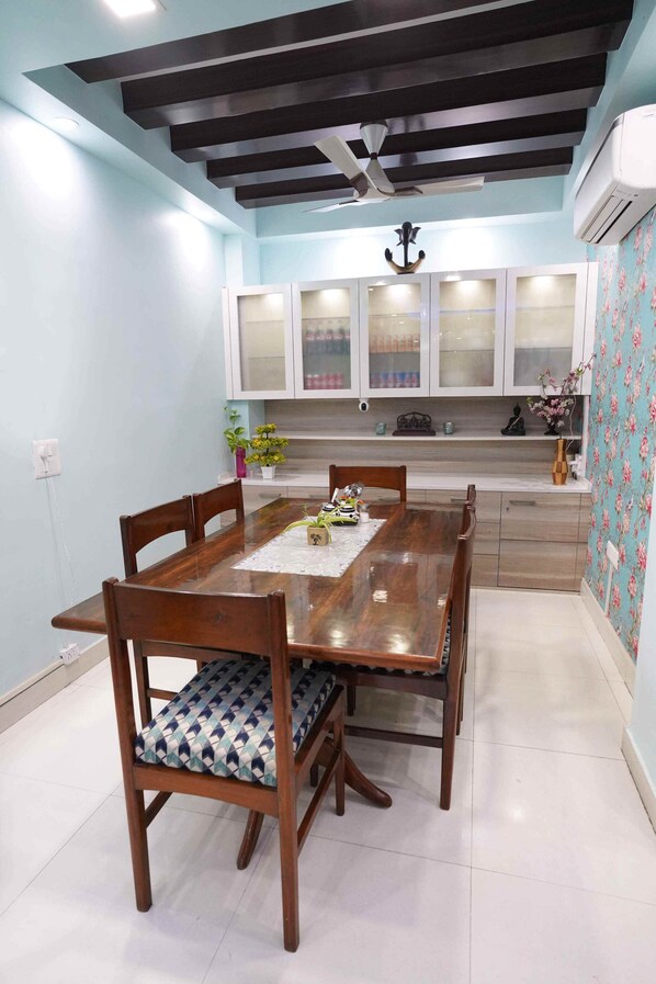 Uma sala de jantar com mesa e cadeiras de madeira, um ventilador de teto e uma cozinha com armários brancos e painel de fundo padronizado.