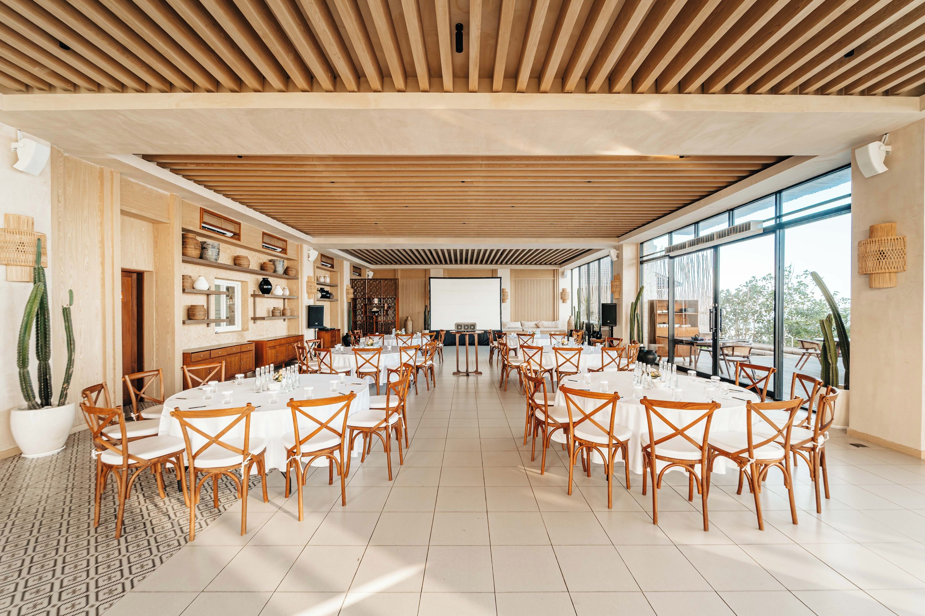 A spacious dining area with round tables and wooden chairs, large windows, and a wooden ceiling.