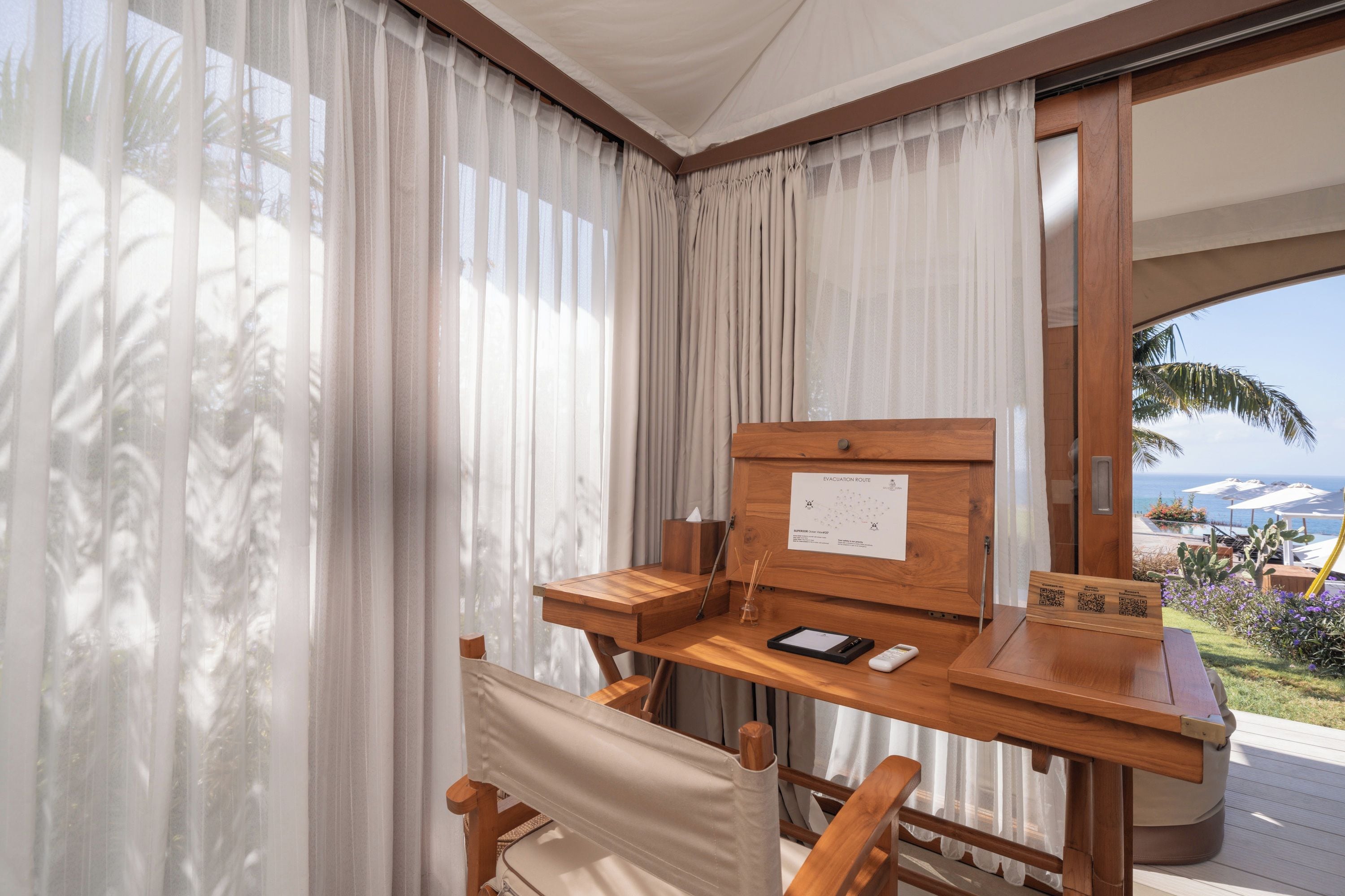 Una habitación con un escritorio de madera, una silla, un ventanal con cortinas traslúcidas y vista al océano.
