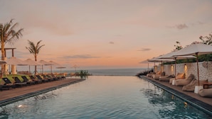 A poolside area with lounge chairs and umbrellas, a stone wall, and a view of the ocean at sunset. - Glamp Nusa (Nusa Dua)