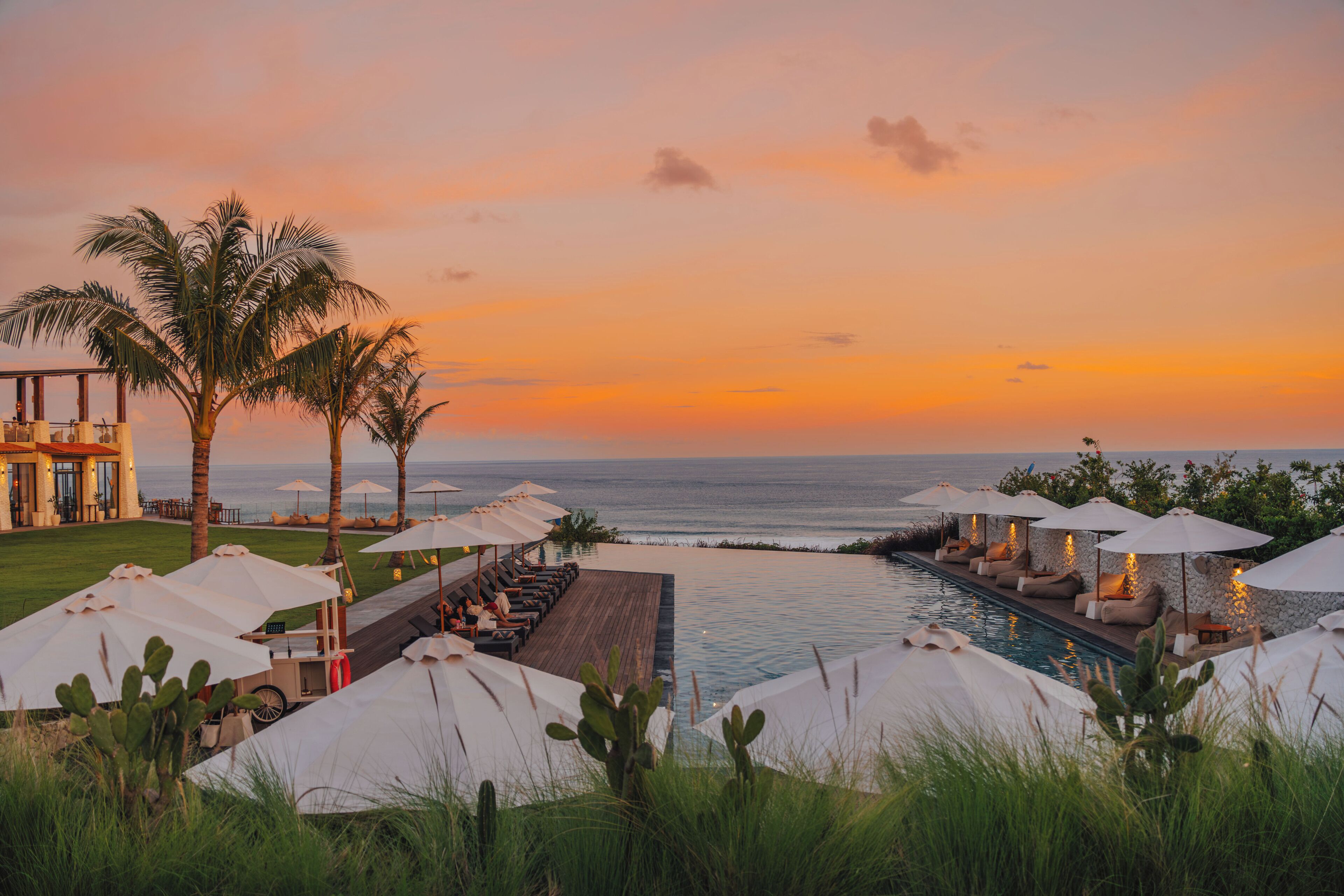 Un espace piscine aménagé dans un complexe hôtelier, avec des chaises longues et des parasols, offrant une vue sur l’océan au coucher du soleil.