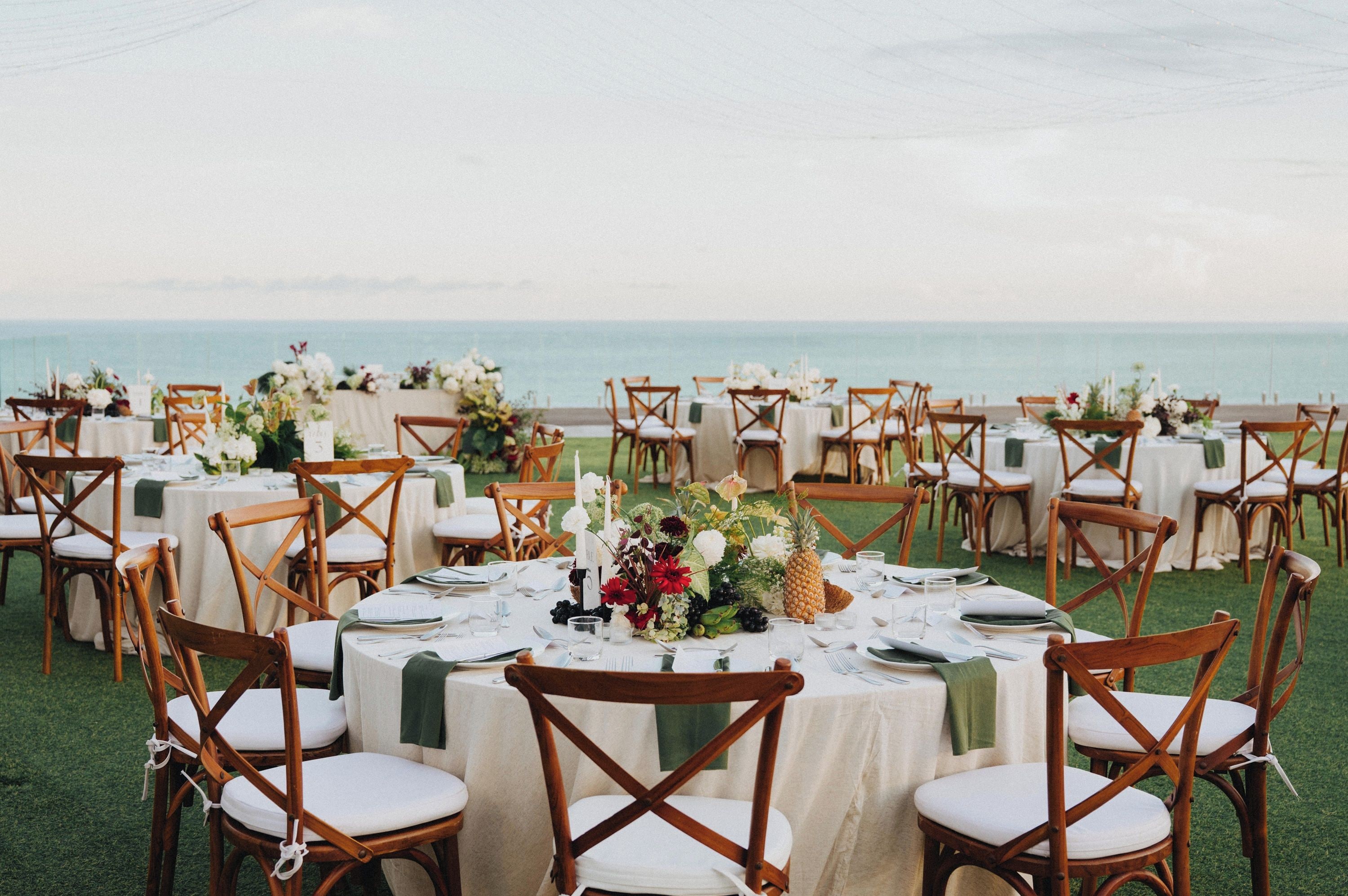 Un espacio de comedor al aire libre con mesas redondas y sillas de madera, dispuesto en un área cubierta de césped con vista al mar.