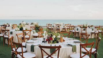 Un espacio de comedor al aire libre con mesas redondas y sillas de madera, dispuesto en un área cubierta de césped con vista al mar.