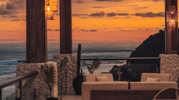 A modern interior with a view of the ocean at sunset, featuring a stone wall, a large vase, and a wooden ceiling. - Glamp Nusa (Nusa Dua)