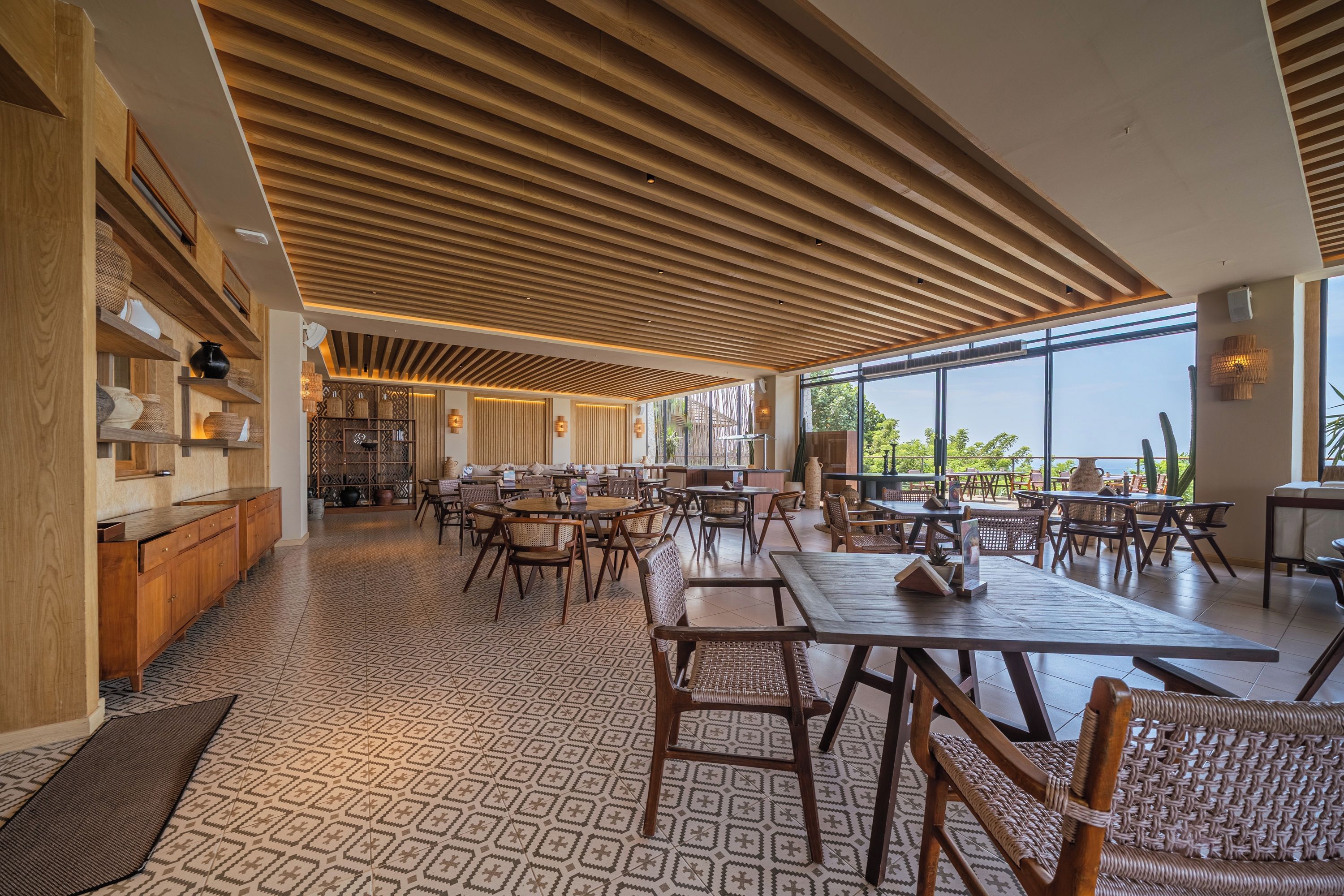 A spacious dining area with wooden tables and chairs, a patterned floor, and large windows offering a view.
