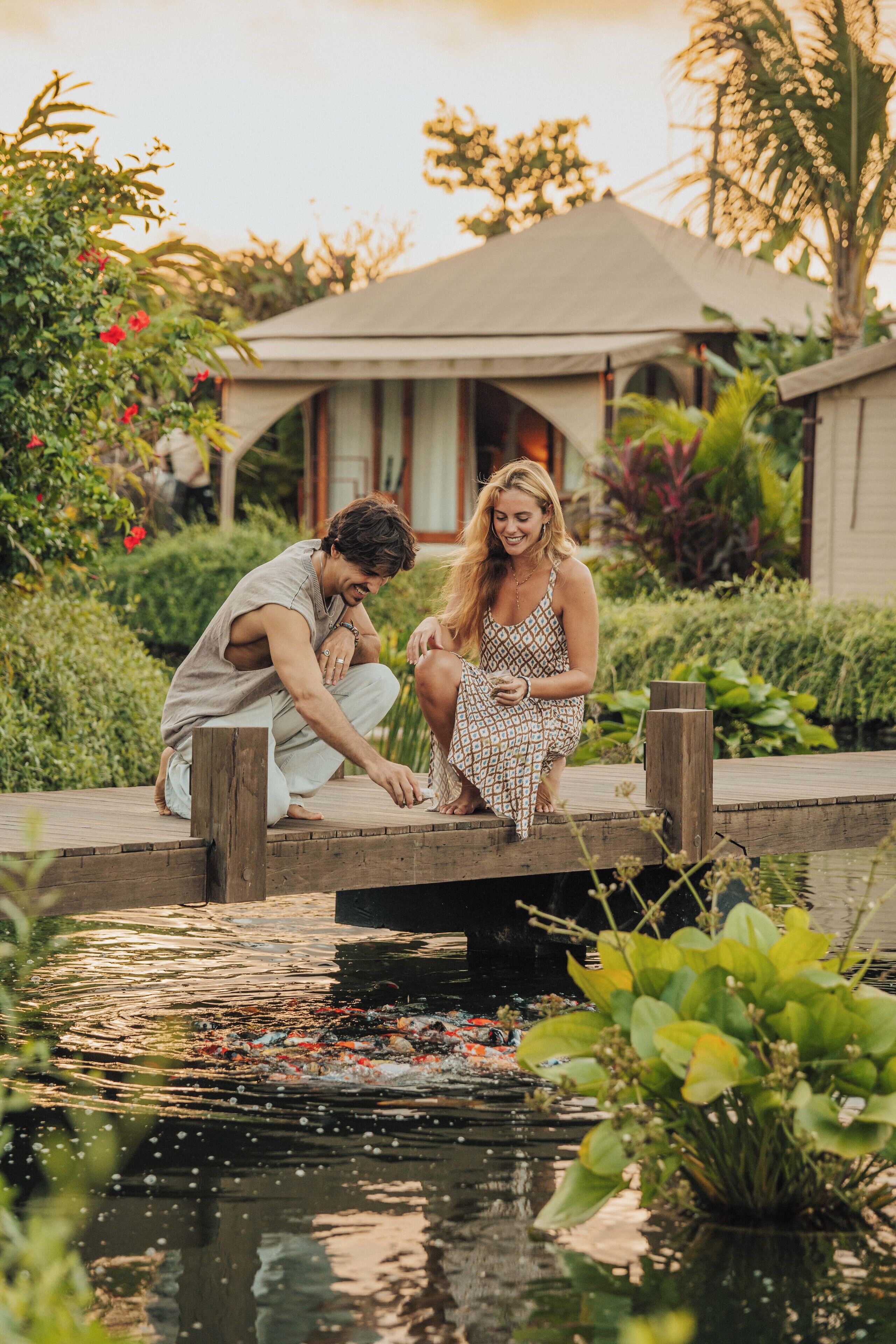 A couple sitting on a wooden dock over a pond, surrounded by lush greenery and a house in the background.
