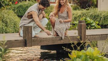 A couple sitting on a wooden dock over a pond, surrounded by lush greenery and a house in the background.