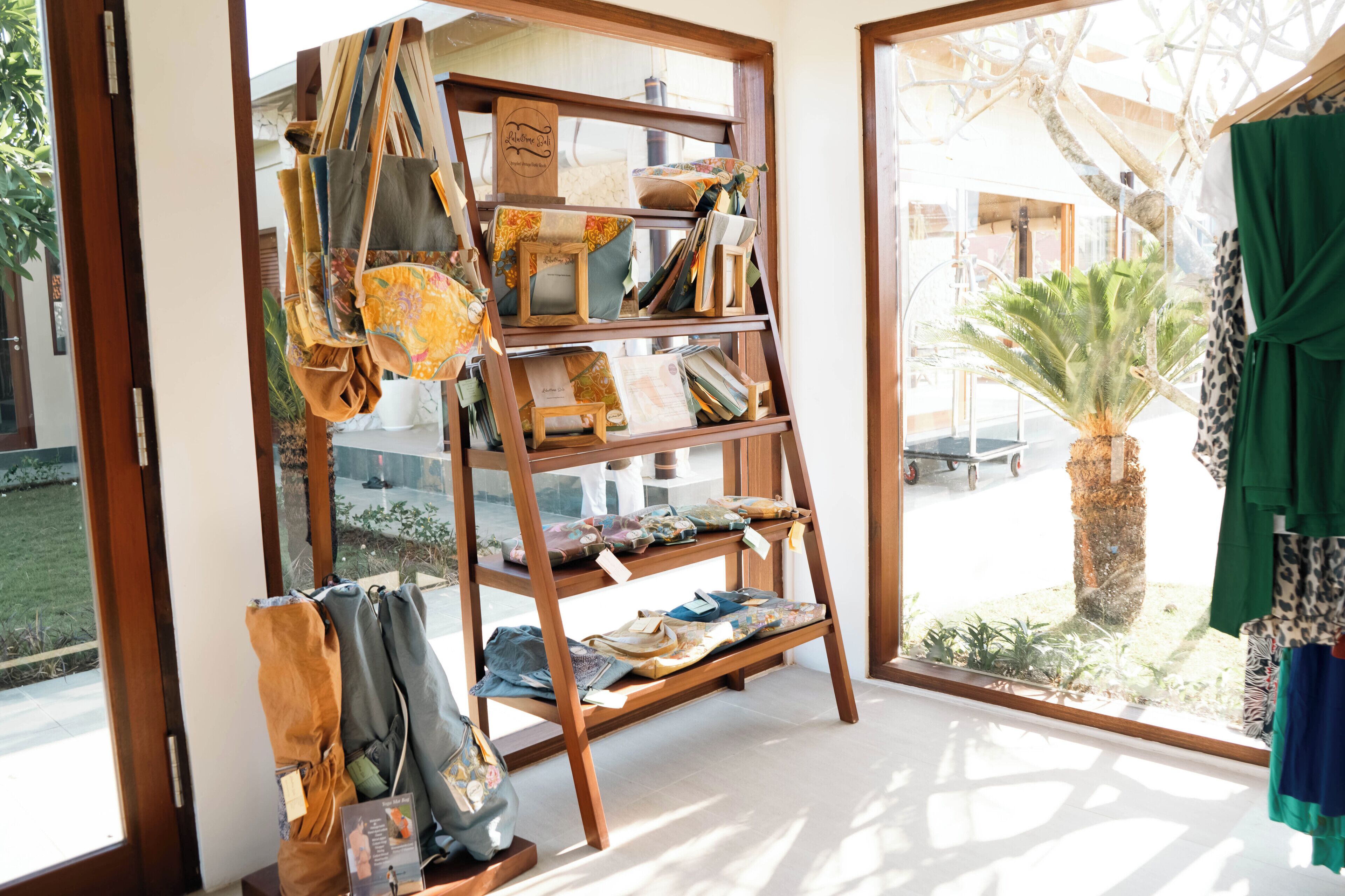 Una estantería de madera con diferentes bolsas y textiles, ubicada cerca de un ventanal con vista a una palmera y un edificio.