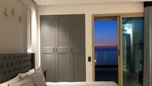 Ein Schlafzimmer mit einem großen Bett, einem grauen Kleiderschrank und einer Glastür, die auf einen Balkon mit Sonnenuntergangsblick führt.