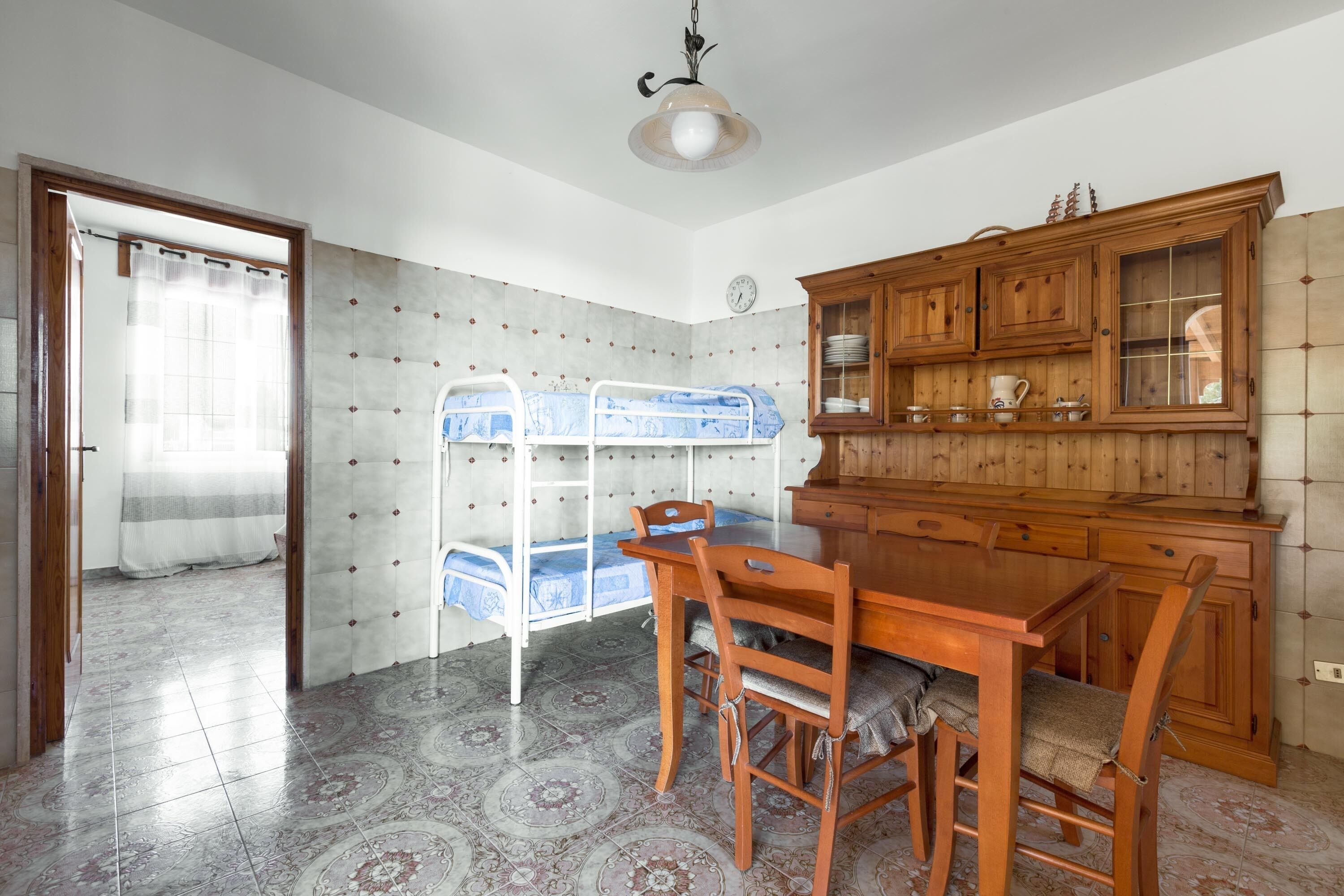 Une pièce avec un lit superposé, une table à manger en bois, des chaises, un buffet et une horloge au mur.