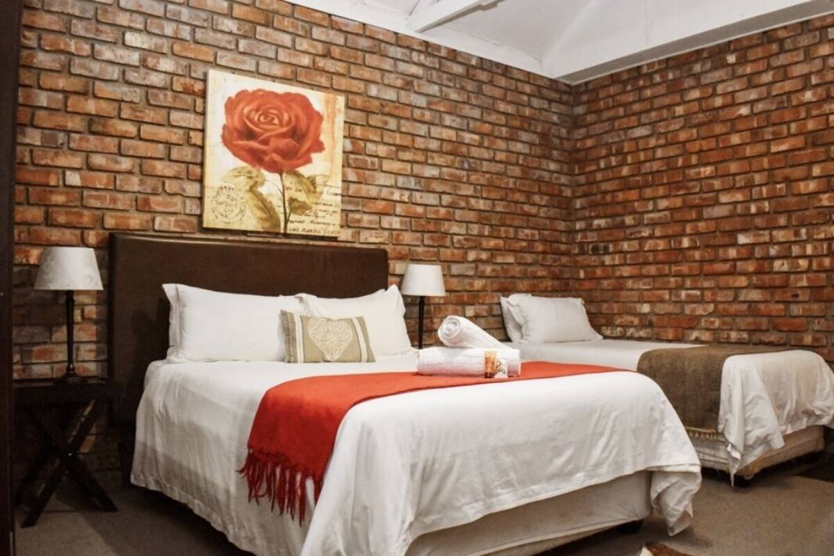 Habitación de hotel con pared de ladrillo, una cama grande con ropa blanca, dos mesitas de noche con lámparas y un cuadro de una rosa roja en la pared.